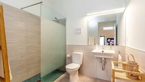 Une salle de bain moderne avec une douche à parois de verre, des toilettes blanches, un lavabo et une étagère en bois où sont posées des serviettes.