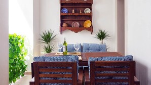 Un coin repas avec une table en bois dressée pour deux personnes, une nappe à motifs et une étagère murale ornée d’assiettes décoratives.