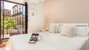 Habitación de hotel con una cama grande, una mesita de noche pequeña, una ventana con vista a un edificio y un balcón con plantas.