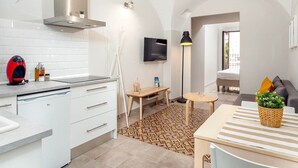 Una cocina moderna con armarios blancos, un hervidor rojo y una estufa empotrada. Hay una pequeña mesa de madera con una lámpara y un televisor montado en la pared.
