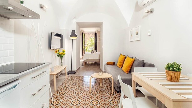 Una cocina moderna con armarios blancos y encimera negra, un comedor con mesa y sillas de madera, y una sala de estar con sofá y cojines.