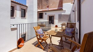 Un patio exterior iluminado por el sol con una mesa y sillas de madera, una maceta naranja pequeña y una pared blanca con una ventana.