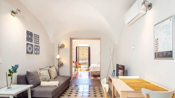 A modern living room with a sofa, a dining table with chairs, a coffee table, and a TV. There is a patterned rug on the floor and a wall-mounted air conditioner.