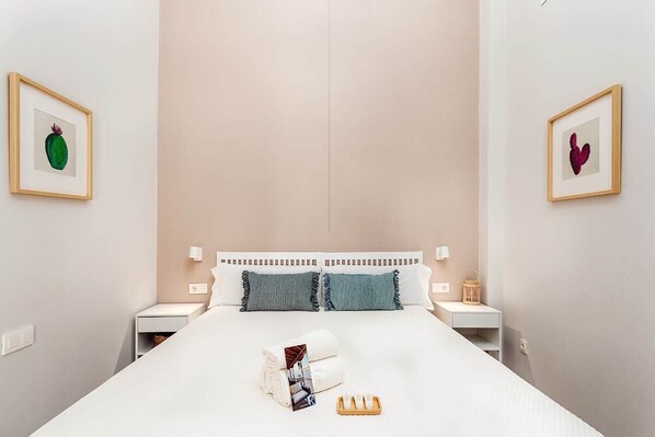 Una cama bien hecha con edredón blanco, dos almohadas, una mesita de noche con lámpara y una obra de arte con un cactus enmarcado en la pared.