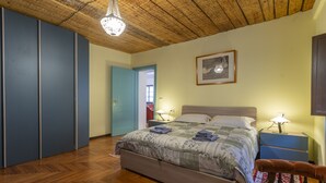 A bedroom with a wooden floor, a bed with a quilt, bedside lamps, a wooden chair, and a door leading to another room. - Hladik House - Alpi Giulie Cosy Apartment (Tarvisio)