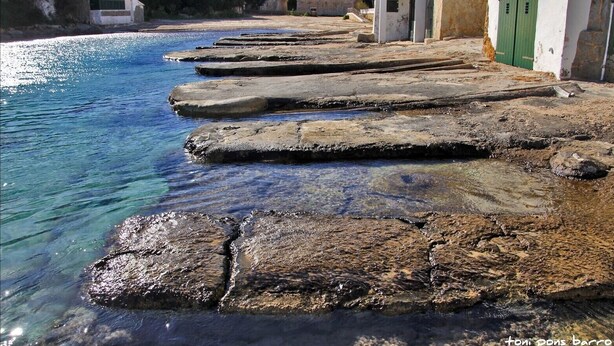 A coastal area with a series of stone steps leading into clear blue water, a white building with green doors, and a stone structure in the background. - Maison de Pécheurs à 1mn à Pied de la Plage Cala Alcaufar (Alcaufar)