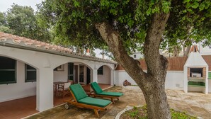 A covered outdoor area with two green lounge chairs, a large tree, and a white building with a tiled roof. - Maison de Pécheurs à 1mn à Pied de la Plage Cala Alcaufar (Alcaufar)