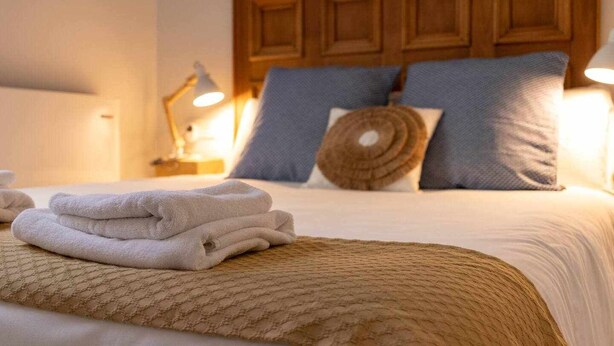A neatly made bed with a beige blanket, a stack of white towels, and a wooden headboard with multiple pillows. - KUIA apartment in Barakaldo, next to the metro (Barakaldo)