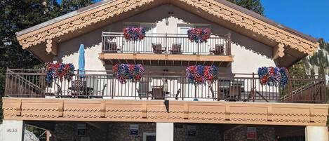 A two-story building with a stone facade, a balcony with flower baskets, and a parking area with two cars.