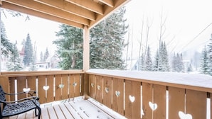 Een houten terras met een bankje, omringd door met sneeuw bedekte bomen en een helder uitzicht op een huis op de achtergrond.