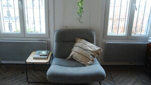 Ein Zimmer mit einem grauen Sessel, einem kleinen Holztisch mit Büchern und einer hängenden Pflanze.