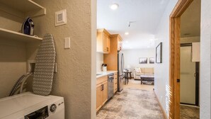 A laundry area with a washing machine and an ironing board, a kitchen with wooden cabinets and a refrigerator, and a living area with a sofa and a coffee table.