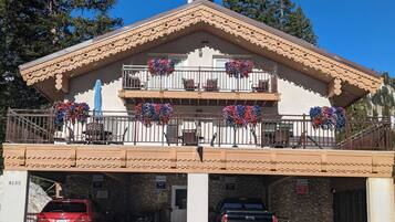 A two-story building with a stone facade, a balcony with flower baskets, and a parking area with two cars.