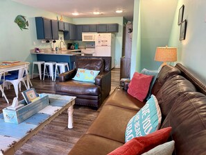 A living room with a brown leather sofa, a coffee table, a chair with a cushion, a kitchen area with cabinets and appliances, and a dining area with stools.