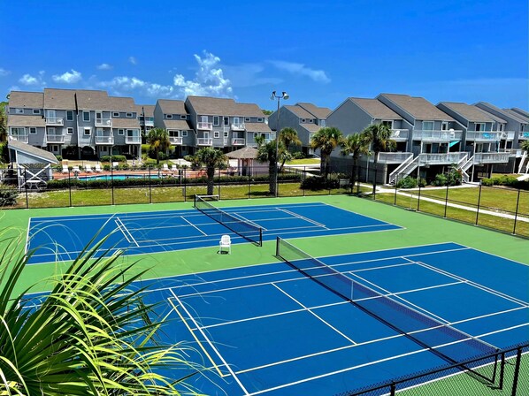 A tennis court with a net, surrounded by a fence, and residential buildings in the background.