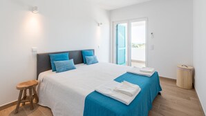 A bedroom with a large bed, two pillows, a stool, a waste bin, and a window with blinds. - Prainha Luxury Apartment (Alvor)