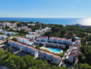 A coastal residential area with white buildings, a central swimming pool, and a view of the sea.