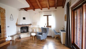 Una sala de estar con chimenea, suelo de madera, un sillón azul, una mesita redonda y una ventana con cortinas.