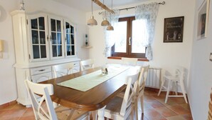 Un comedor con una mesa de madera, sillas blancas, un armario blanco y una ventana con cortinas.
