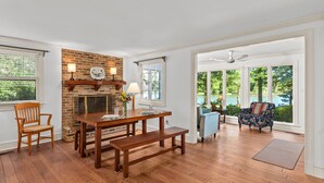 Un vaste salon doté d’un plancher en bois, d’une cheminée en briques, d’une table à manger avec des bancs et de grandes fenêtres offrant une vue sur un lac.