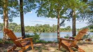 Deux fauteuils Adirondack en bois, placés face à un lac, avec des arbres en arrière-plan et un ciel dégagé.