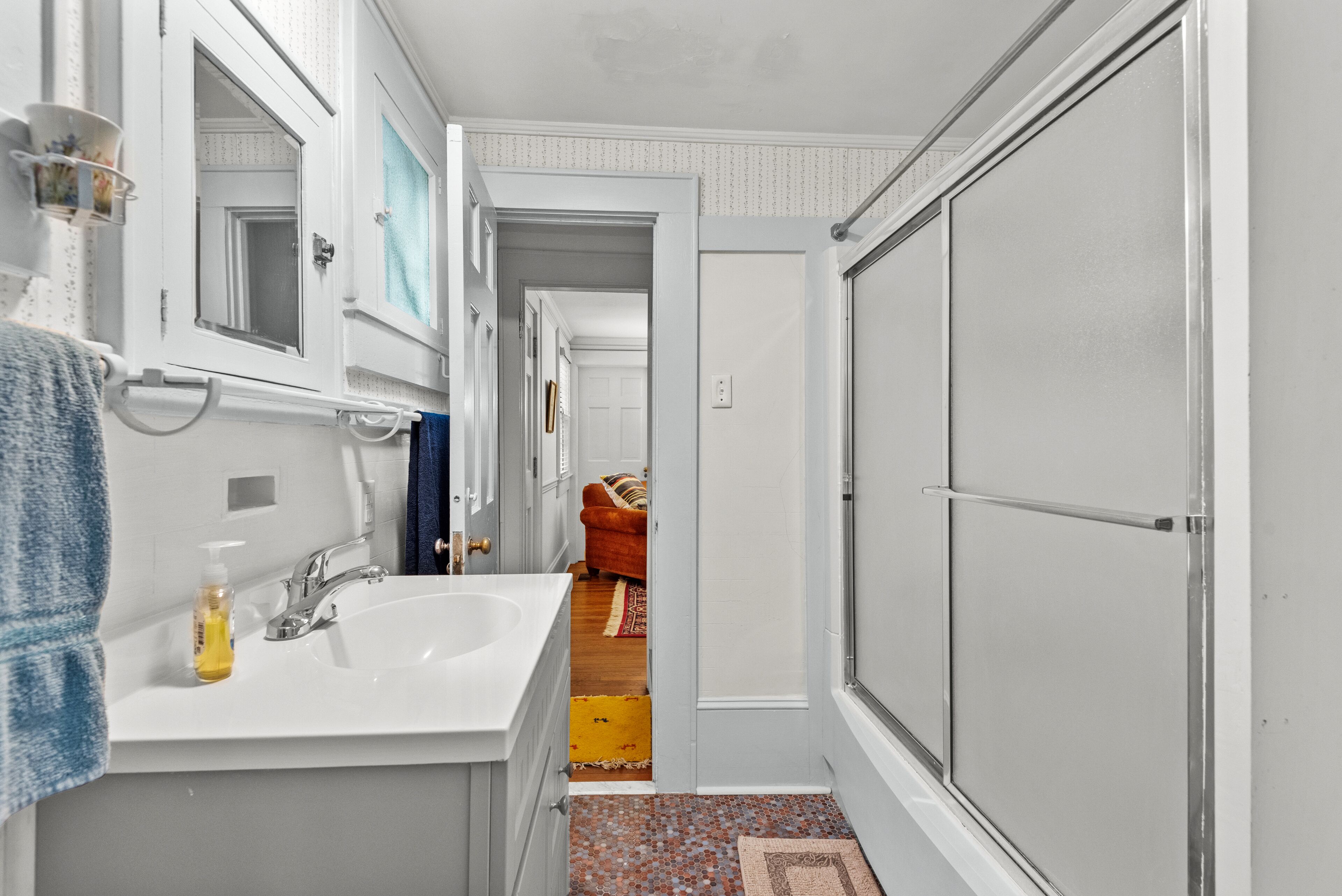 A narrow bathroom with a white sink, mirror, and a shower door. A blue towel hangs on the wall, and a yellow bottle is on the sink.