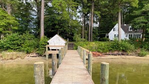Une jetée en bois mène à une petite construction en bois à la façade blanche, entourée d’une végétation luxuriante et avec une maison en arrière-plan.
