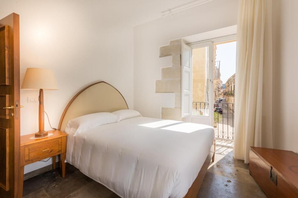 Un dormitorio con cama, mesita de noche, lámpara y vista a la calle a través de una puerta balcón.
