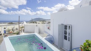 Una piscina en la azotea con agua azul cristalina y pétalos de flores morados, rodeada de edificios blancos y vistas al mar y montañas lejanas.