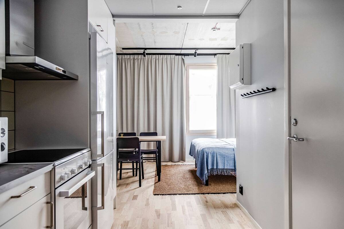 Ett kompakt kök med moderna vitvaror, en matplats med bord och stolar, en säng med ett blått överkast och ett stort fönster med gardiner.
