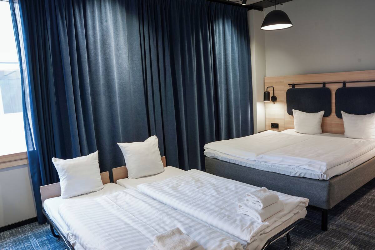 Ett hotellrum med två sängar, mörka gardiner, en sänggavel i trä och en sänglampa.