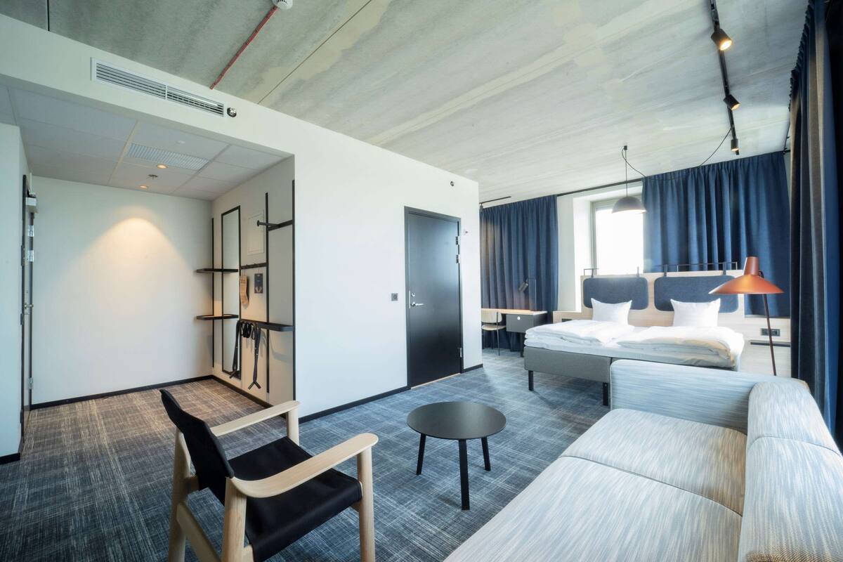 Ett modernt hotellrum med en stor säng, en soffa, ett litet bord och ett skrivbord. Det finns en garderob, en spegel och ett fönster med gardiner.