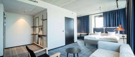 A modern hotel room with a large bed, a sofa, a small table, and a desk. There is a wardrobe, a mirror, and a window with curtains.
