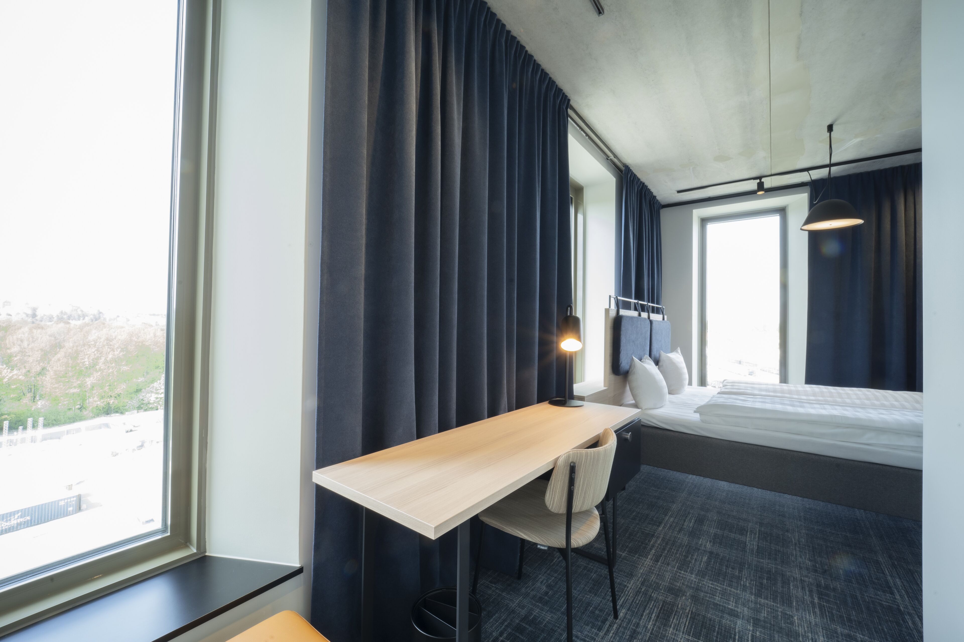 Habitación de hotel con cama, escritorio, silla y un amplio ventanal con cortinas.