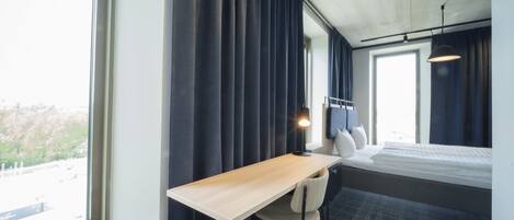 Habitación de hotel con cama, escritorio, silla y un amplio ventanal con cortinas.