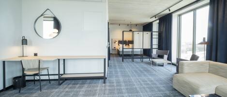 Quarto de hotel moderno com escrivaninha, cadeira, sofá e janelas amplas.