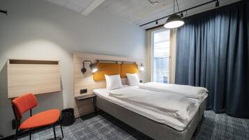 A hotel room with a bed, a desk, an orange chair, and a window with curtains.