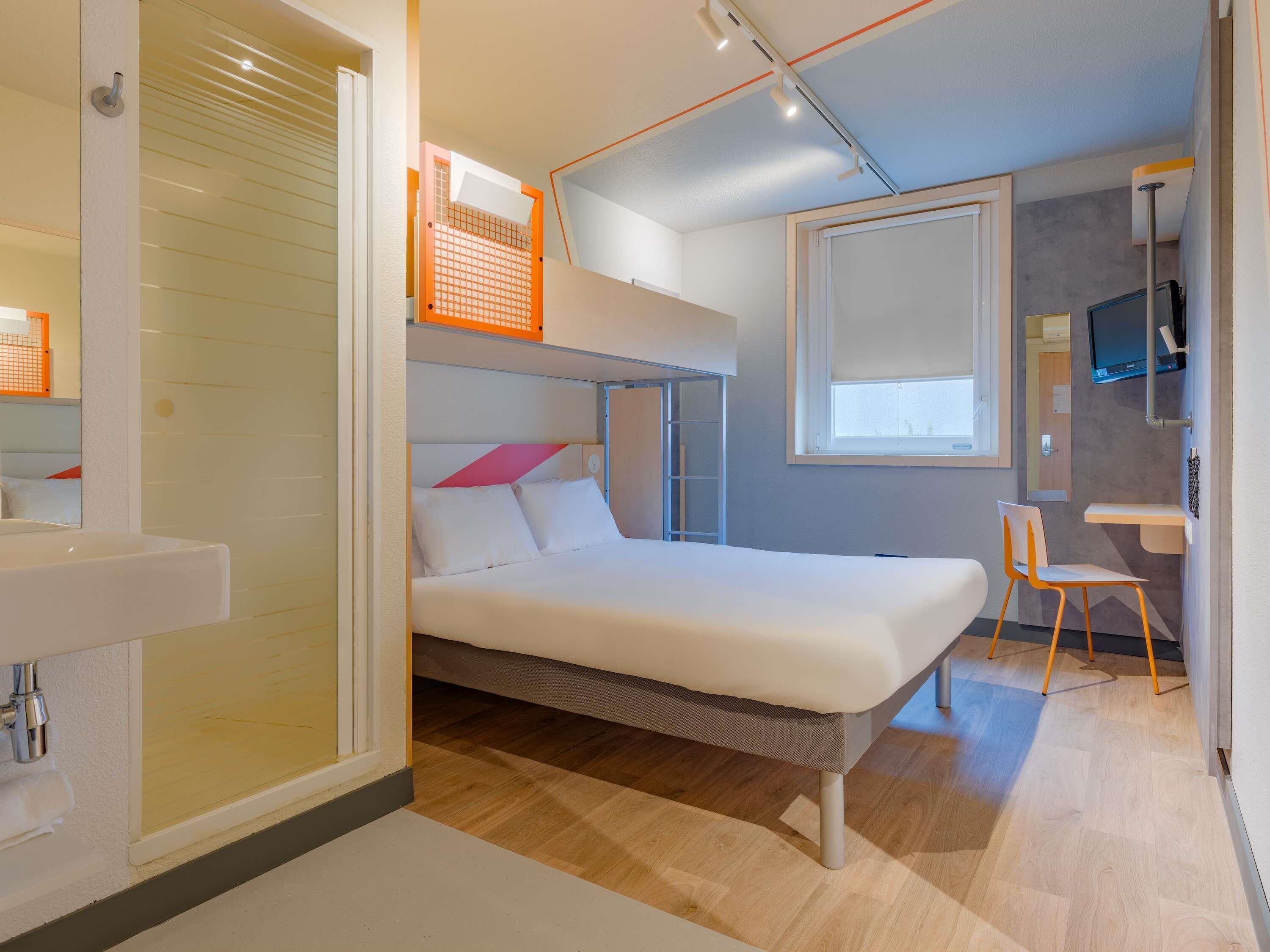 A compact hotel room with a bunk bed, a small desk, a chair, a TV, and a bathroom with a sink.