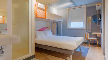 A compact hotel room with a bunk bed, a small desk, a chair, a TV, and a bathroom with a sink.