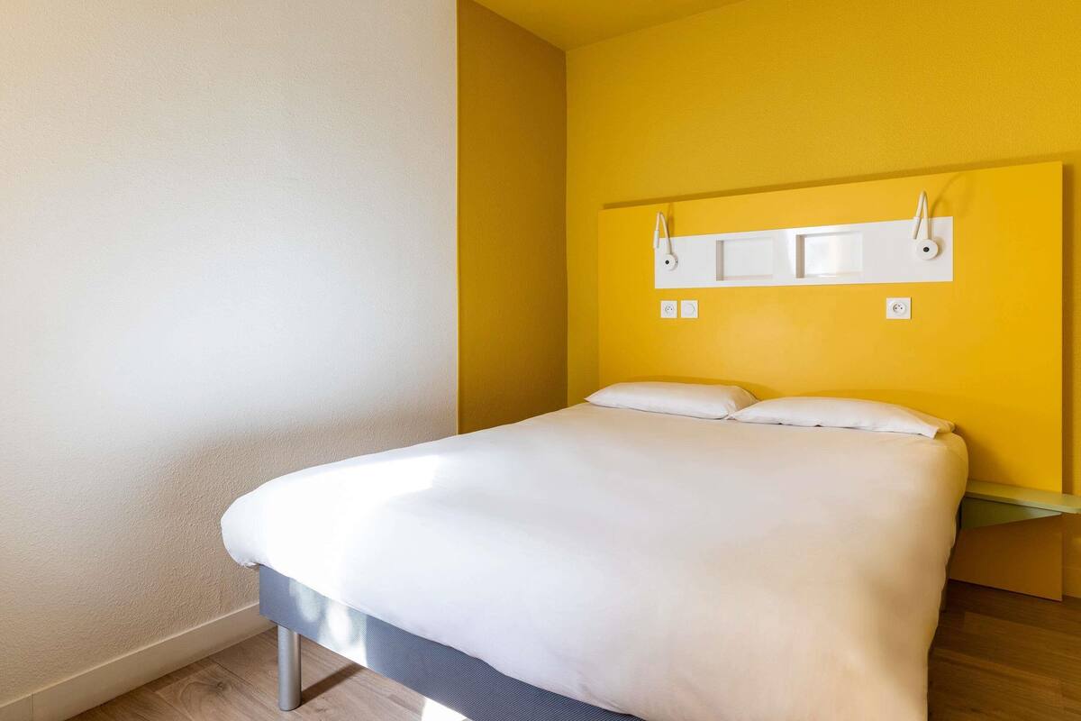 Una habitación de hotel con una cama, cabecera con lámpara y panel de control en la pared.