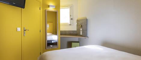 A hotel room with a bed, a television mounted on a yellow wall, a small window, and a wooden shelf.