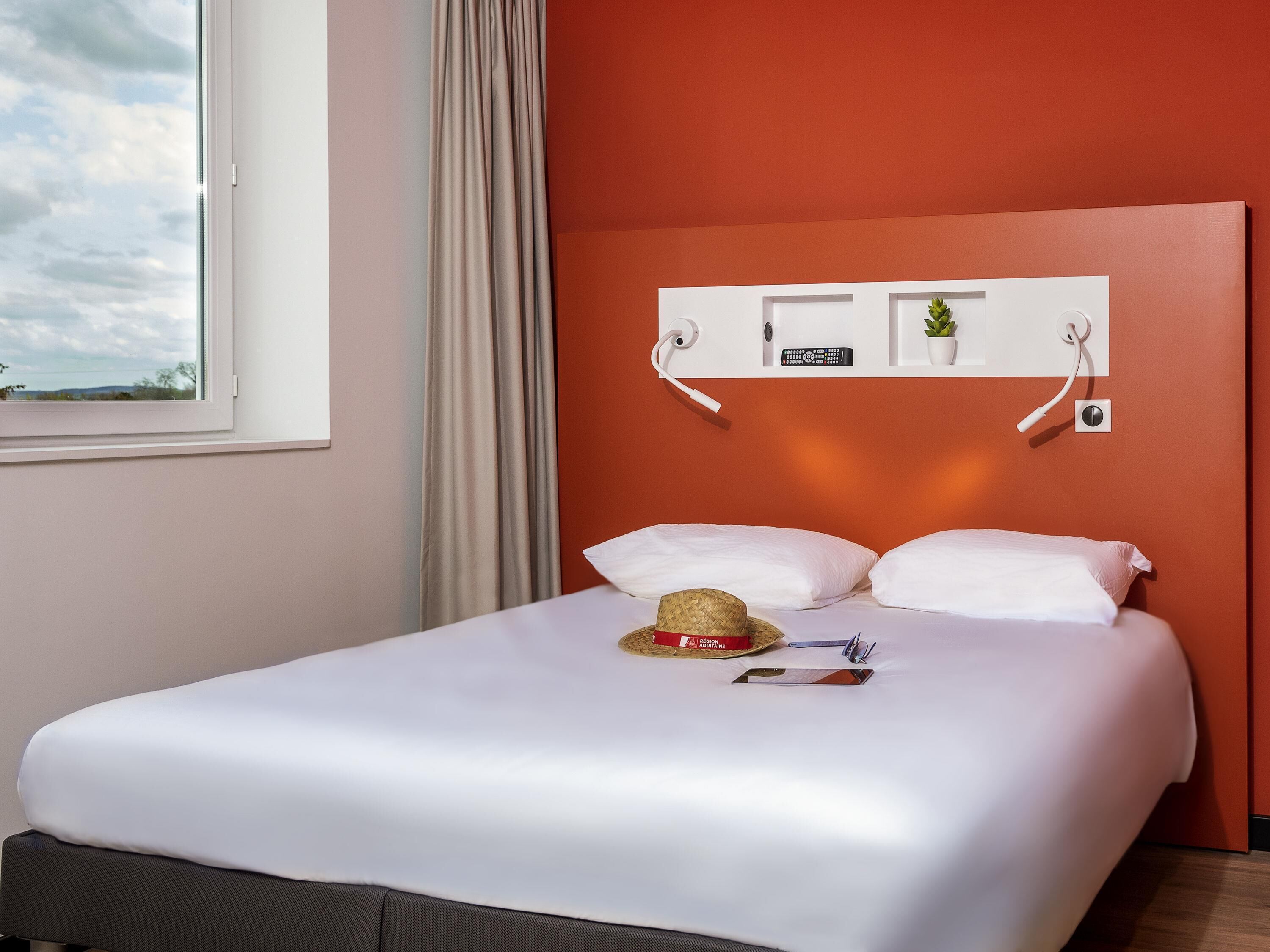 Habitación de hotel con cama, ventana, cortina, televisor de pared y toma de corriente.