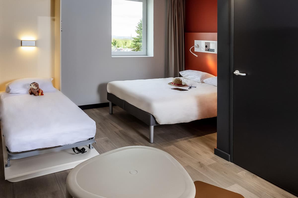 Quarto de hotel com duas camas, uma mesa pequena e uma cadeira. Há uma janela com vista para o verde lá fora.