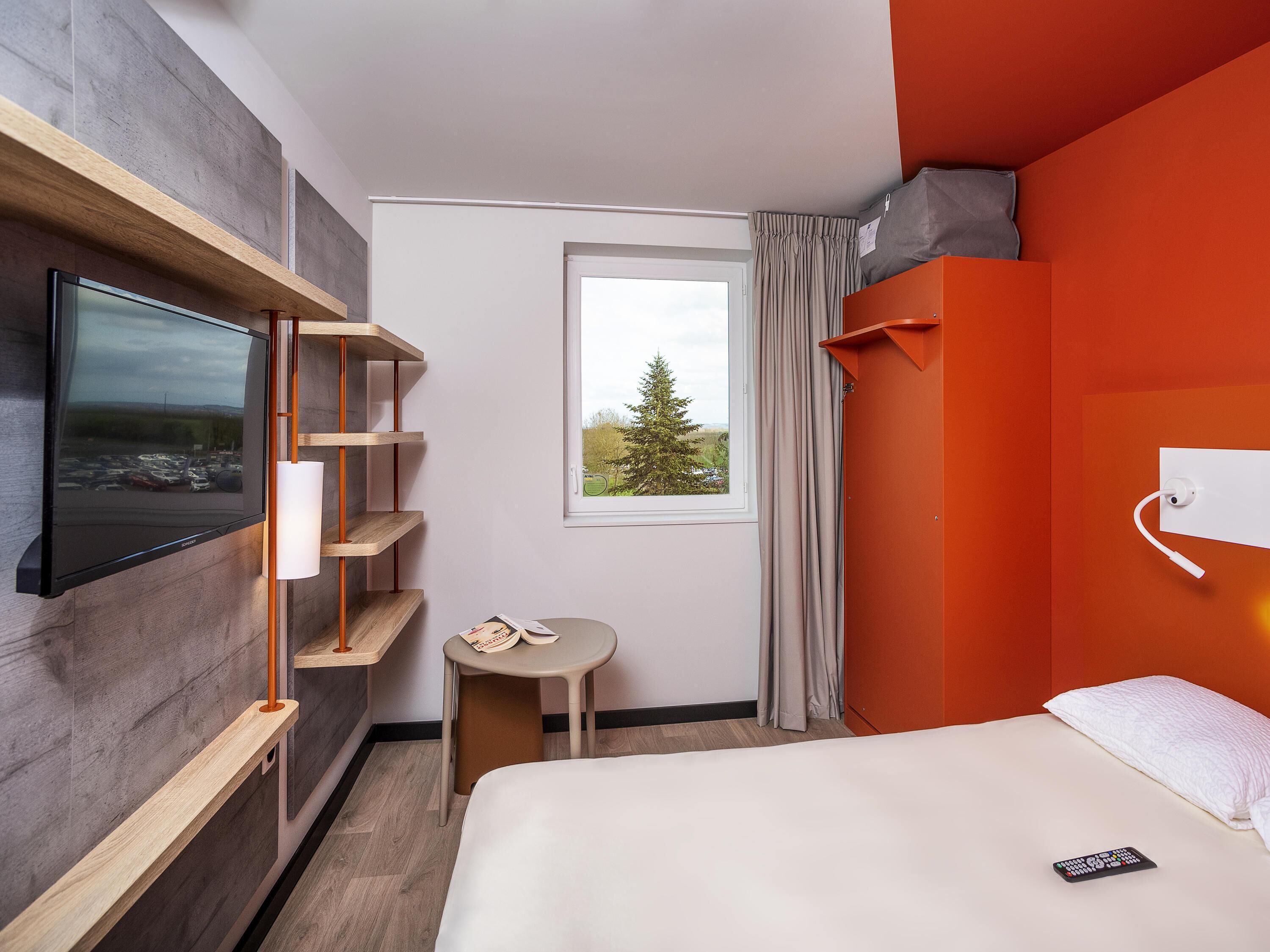 A hotel room with a bed, a TV mounted on the wall, a small table, and a window with curtains.