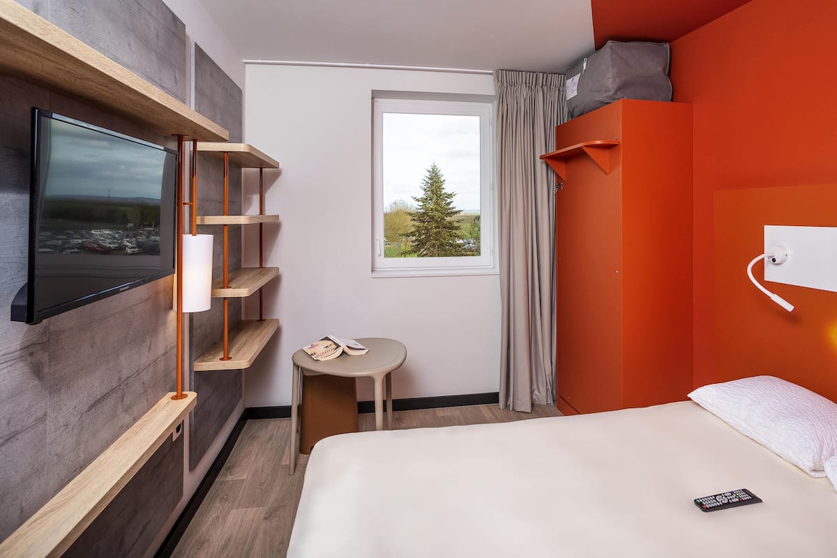 Ein Hotelzimmer mit Bett, Wandfernseher, kleinem Tisch und einem Fenster mit Vorhängen.