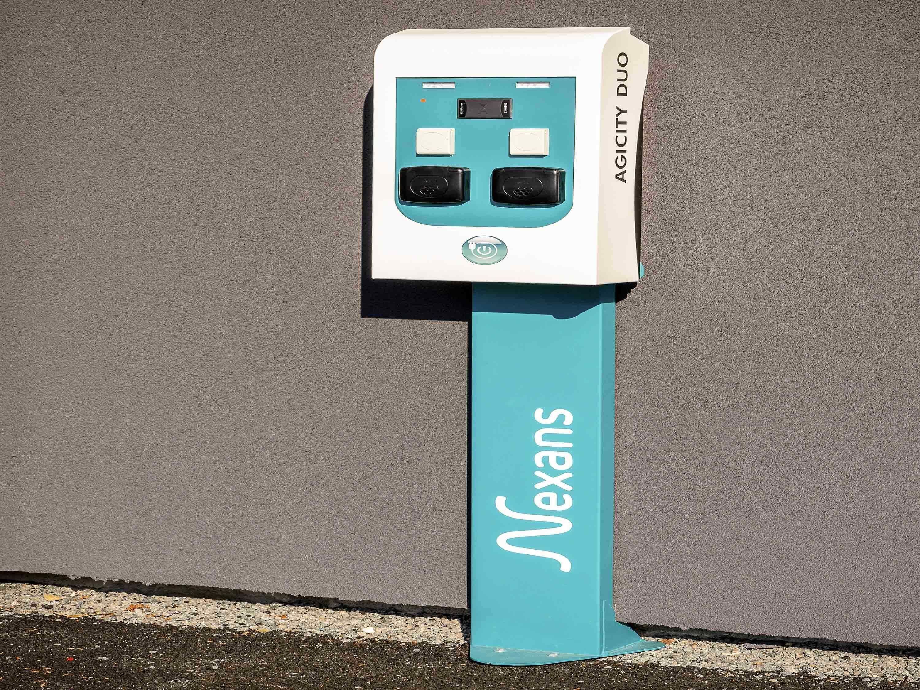 A Nexans branded electrical box with two sockets, mounted on a teal-colored pole against a grey wall.
