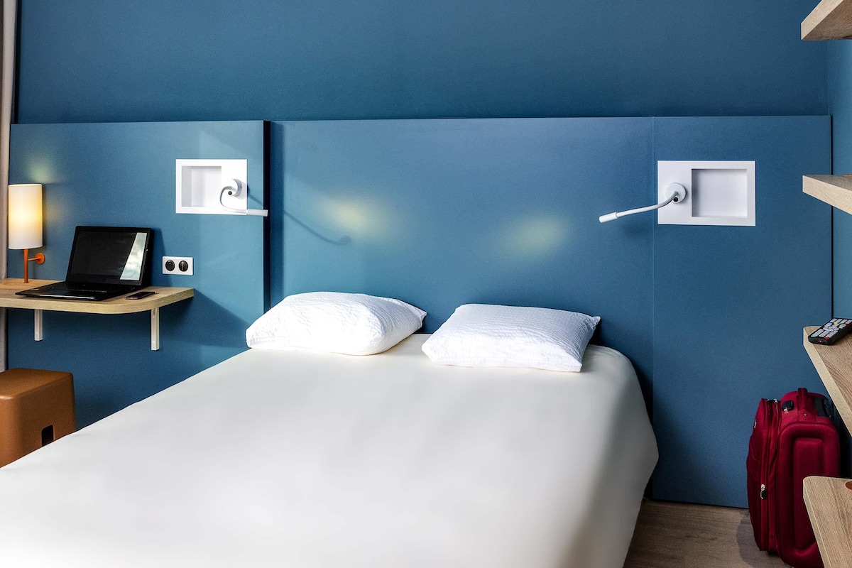 Ein Hotelzimmer mit einem Bett, einem Schreibtisch mit einem Laptop, einer Lampe, einem Telefon und einem roten Koffer.