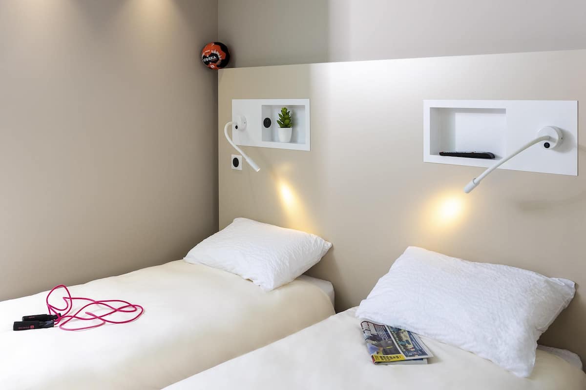 Quarto de hotel com duas camas, cada uma com um travesseiro branco e um cobertor branco. Há prateleiras fixadas na parede com uma planta e um controle remoto. Um cabo de carregamento vermelho está sobre a cama à esquerda.