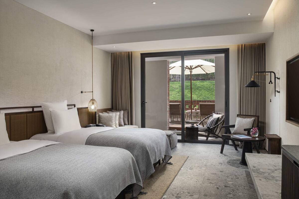 A hotel room with two beds, a small table with chairs, a view of a garden through a glass door, and a large window with curtains.