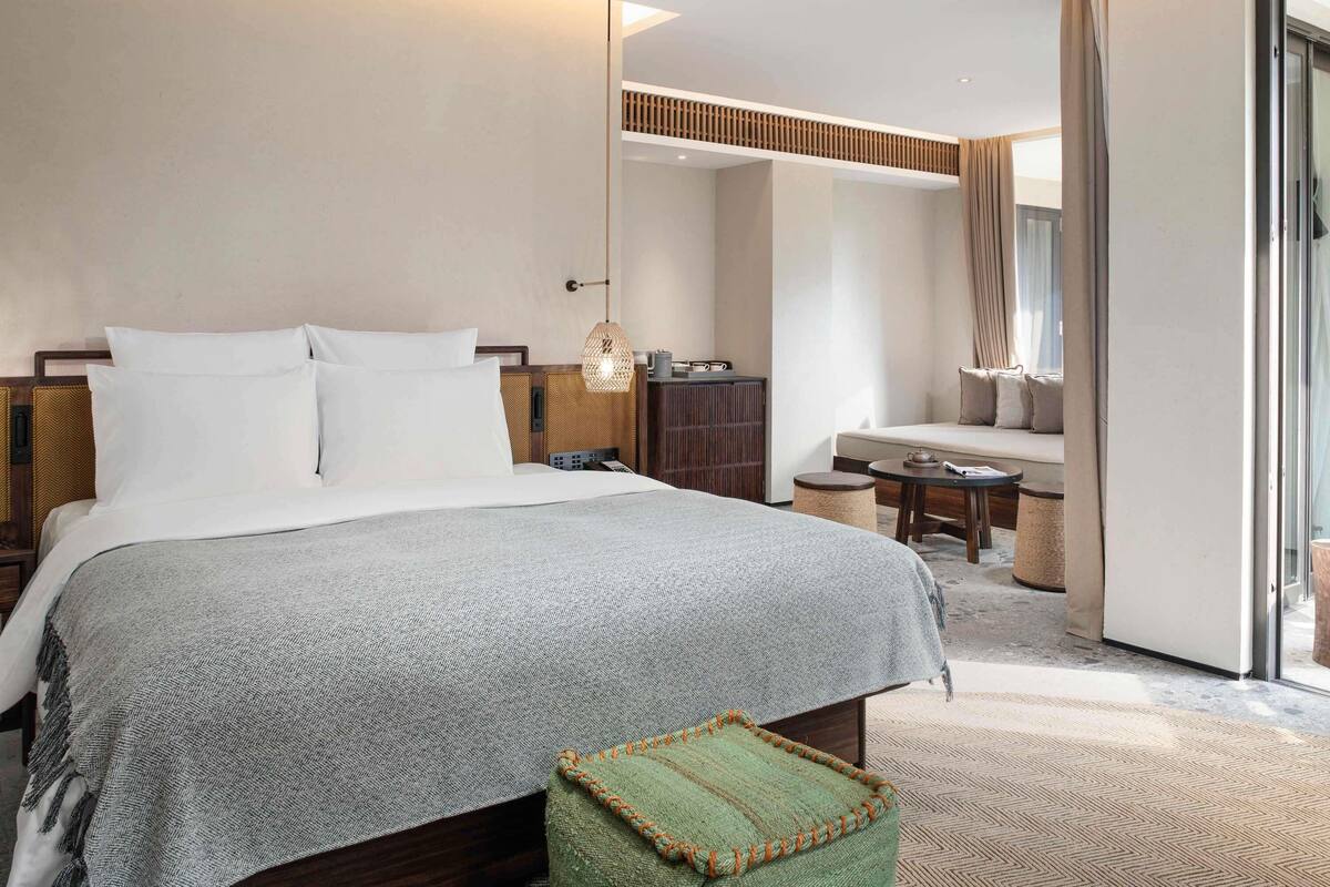 A hotel room with a large bed, a small table, a sofa, and a sliding glass door.